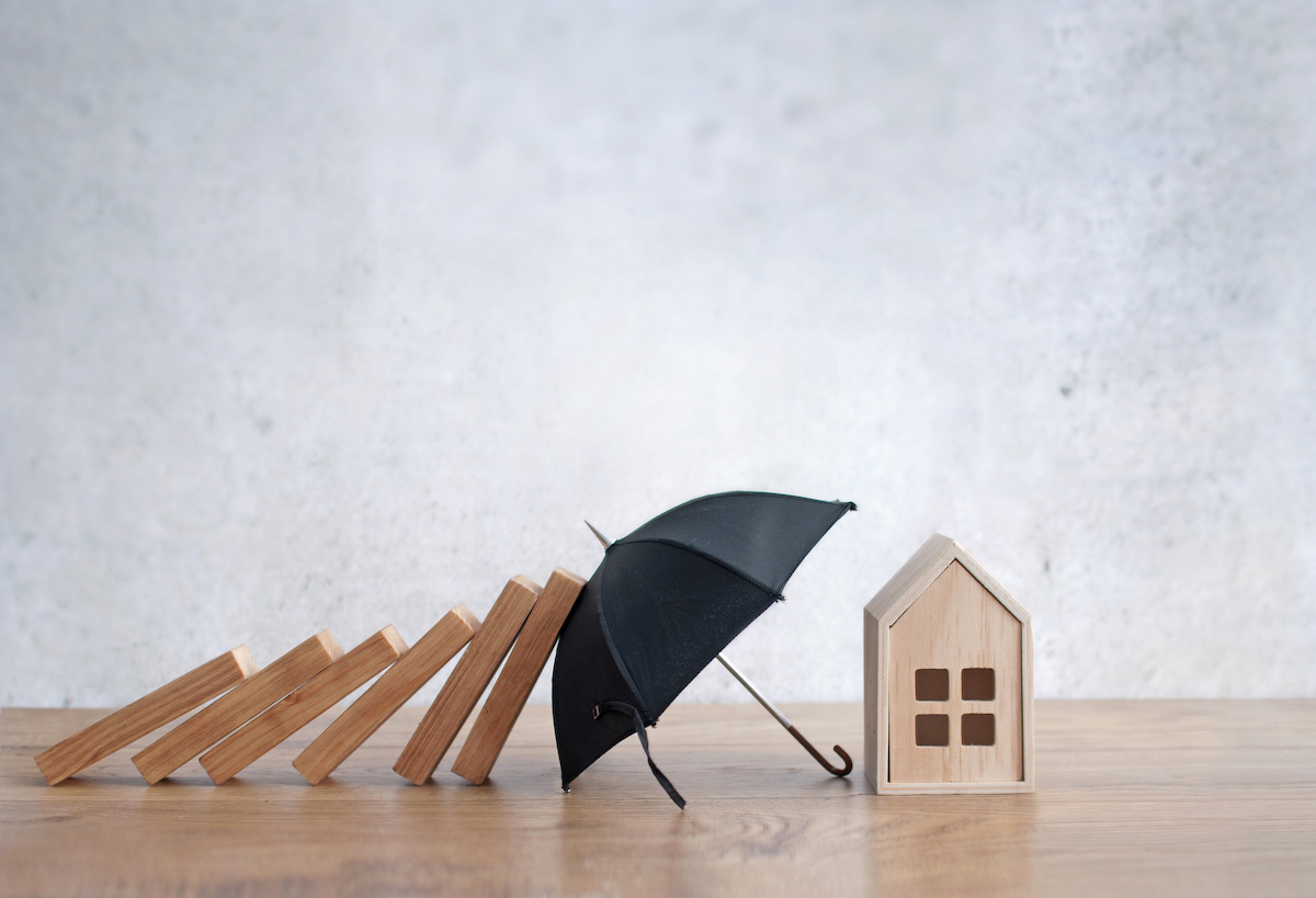 The same way hazard insurance protects your home from perils, in this image a small umbrella blocks a series of blocks falling like dominos from hitting a small toy house.